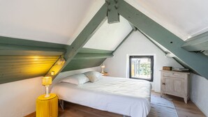 Et soveværelse med skråvæg, en seng med hvide sengetæger, en gul lampe og et lille vindue.