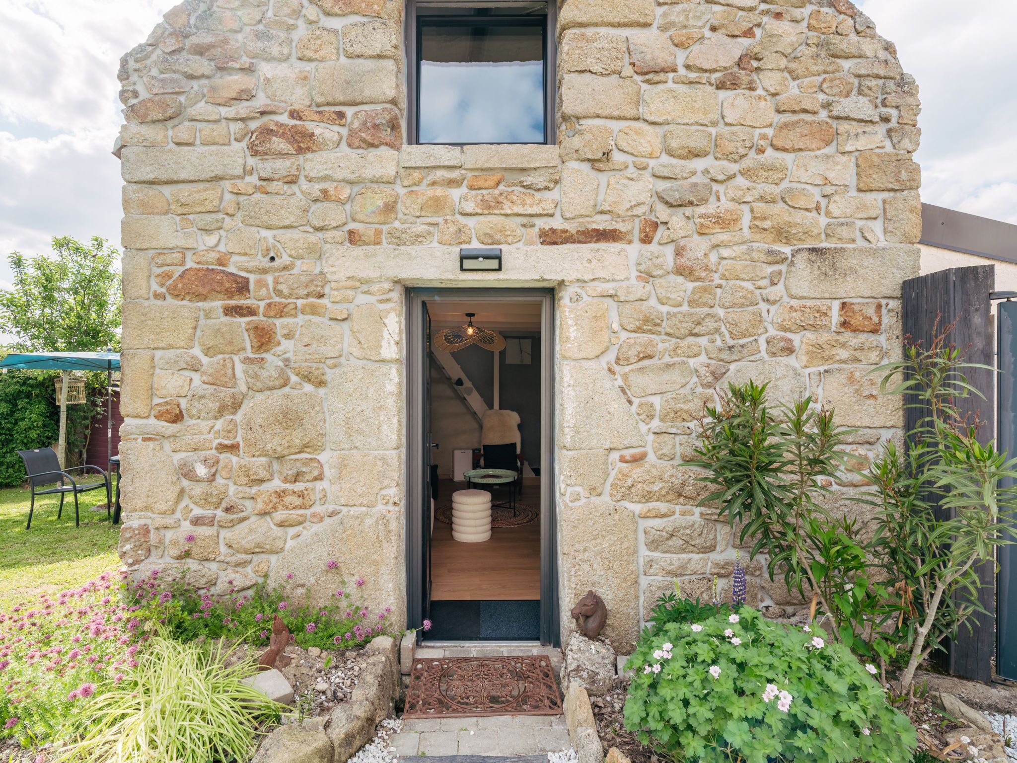 A stone building with a small entrance, a window above the door, and a garden with various plants and flowers.