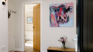 A hallway with a wooden door, a wall-mounted abstract painting, a small wooden table with a vase of flowers, and a coat rack with hooks. - Stables Cottage, Nethybeodge (Nethybridge)