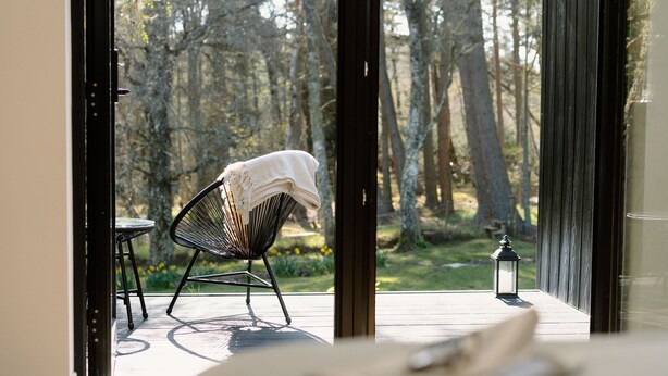 A room with a large window overlooking a forested area, a chair with a white cloth, and a small table with a candle. - Stables Cottage, Nethybeodge (Nethybridge)