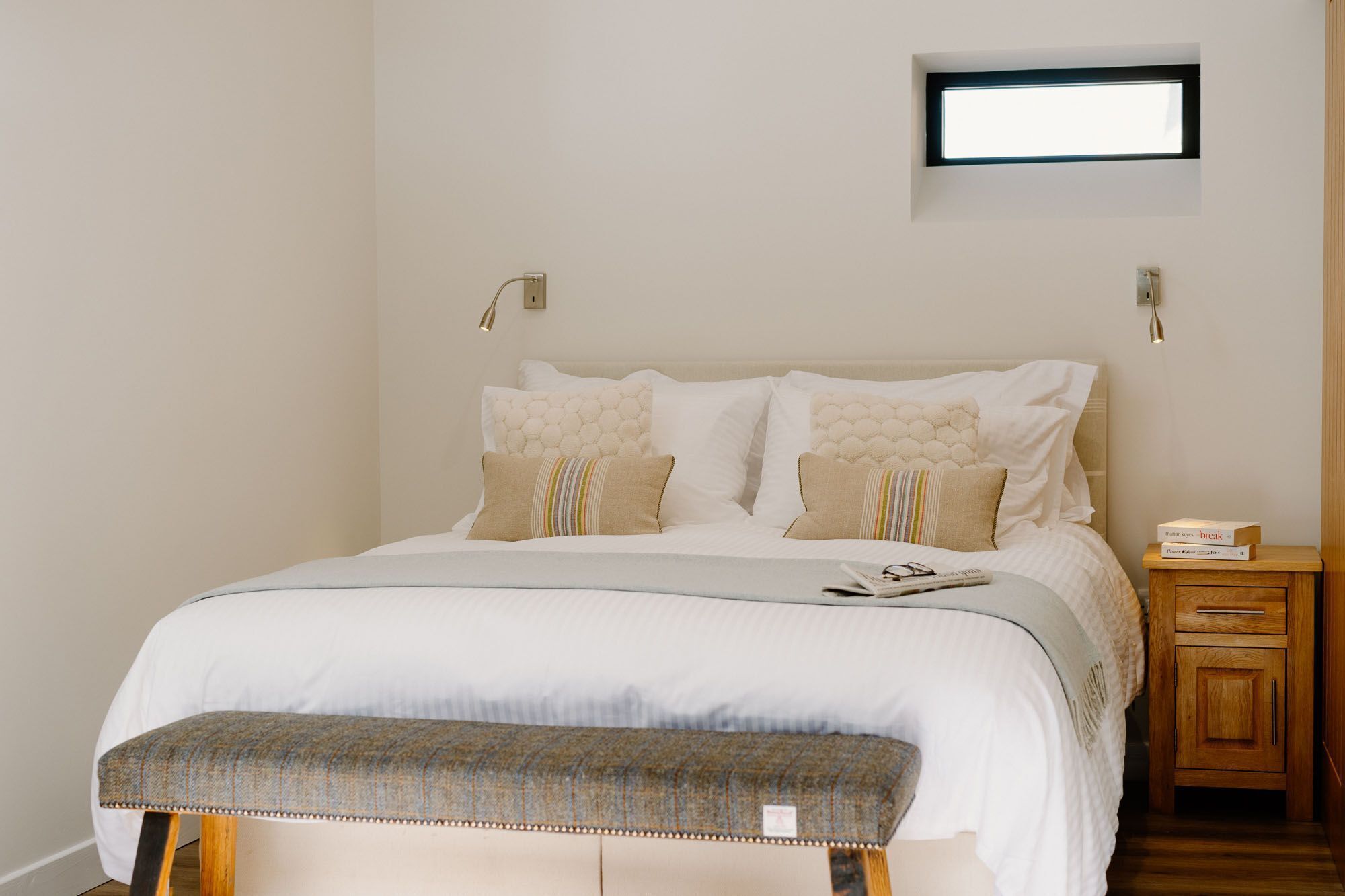 A neatly made bed with a grey bench, a bedside table with books, and a small window above the bed.