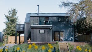 A modern two-story house with a balcony, a chimney, and a small garden with yellow flowers. - Stables Cottage, Nethybeodge (Nethybridge)