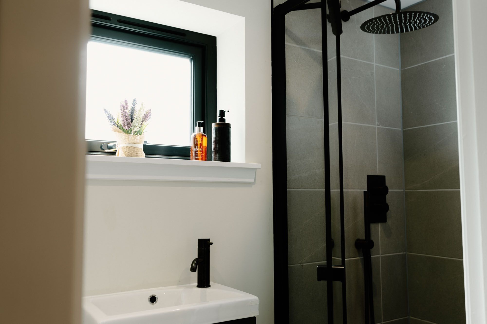 Ein Badezimmer mit einem weißen Waschbecken, einem Fenster darüber und einer Duschkabine mit einer schwarzen Tür.