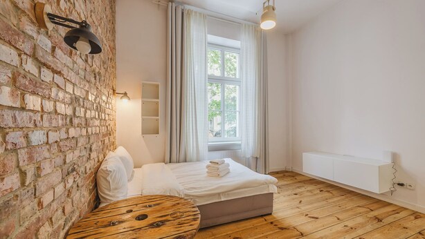 A bedroom with a brick wall, a wooden floor, a bed with white linens, a bedside table, and a window with curtains. - Wilson Park Loft Studio with Mezzanine (Poznań)