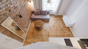 A modern room with a brick wall, wooden flooring, a sofa, a round wooden table, a mirror, and a window with curtains. - Wilson Park Loft Studio with Mezzanine (Poznań)