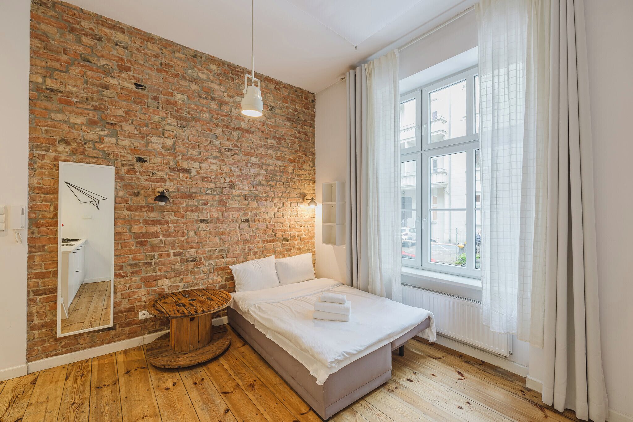 A bedroom with a brick wall, a wooden floor, a bed with white linens, a round wooden stool, a mirror, and a window with curtains.