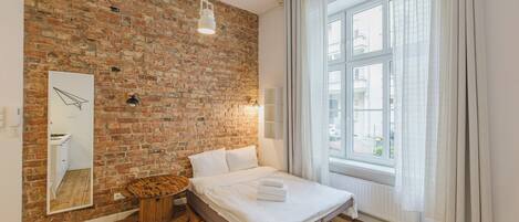 A bedroom with a brick wall, a wooden floor, a bed with white linens, a round wooden stool, a mirror, and a window with curtains.
