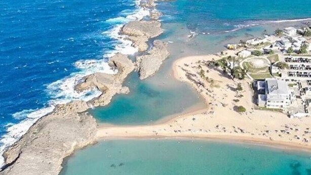 An aerial view of a coastal area with clear turquoise waters, a sandy beach, and a small cluster of buildings.