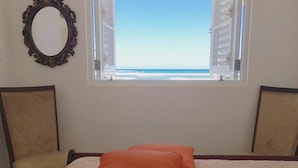 A bedroom with a bed, two chairs, a window with a view of the beach, and a mirror on the wall.