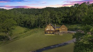 A two-story house with a large deck and a stone fireplace, surrounded by lush greenery and hills. - Stunning Views Hilltop Cabin Near Homestead Resort! (Hot Springs)