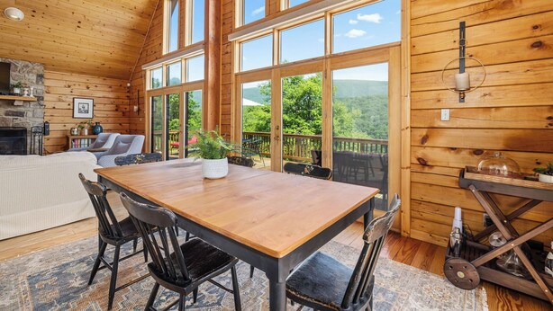 A wooden cabin interior with a dining table, chairs, a fireplace, and large windows offering a view of the outdoors. - Stunning Views Hilltop Cabin Near Homestead Resort! (Hot Springs)