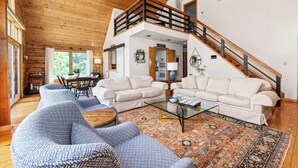 A spacious living room with two beige sofas, a blue patterned sofa, and a glass coffee table. A wooden staircase leads to an upper level. - Stunning Views Hilltop Cabin Near Homestead Resort! (Hot Springs)