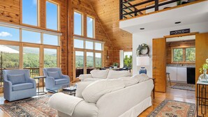 A spacious living room with a large sofa, two armchairs, and a coffee table. The room features large windows offering a scenic view of the mountains. - Stunning Views Hilltop Cabin Near Homestead Resort! (Hot Springs)