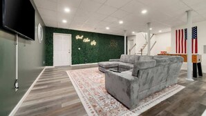 Ein modernes Wohnzimmer mit einem grauen Ecksofa, einem gemusterten Teppich, einem Flachbildfernseher und einer grünen Wand mit einem dekorativen Schild.