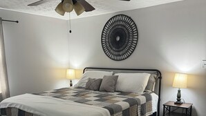 A bedroom with a bed, two bedside lamps, a ceiling fan, a decorative wall piece, and a small nightstand.