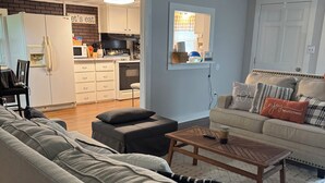 A living room with a sofa, ottoman, and coffee table. There is a kitchen area visible through an open door.