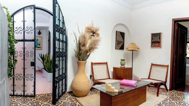 Ein Zimmer mit einem Glastisch, zwei Korbstühlen, einer großen Vase mit Trockenpflanzen, einem schwarzen Tor und einem gefliesten Boden.