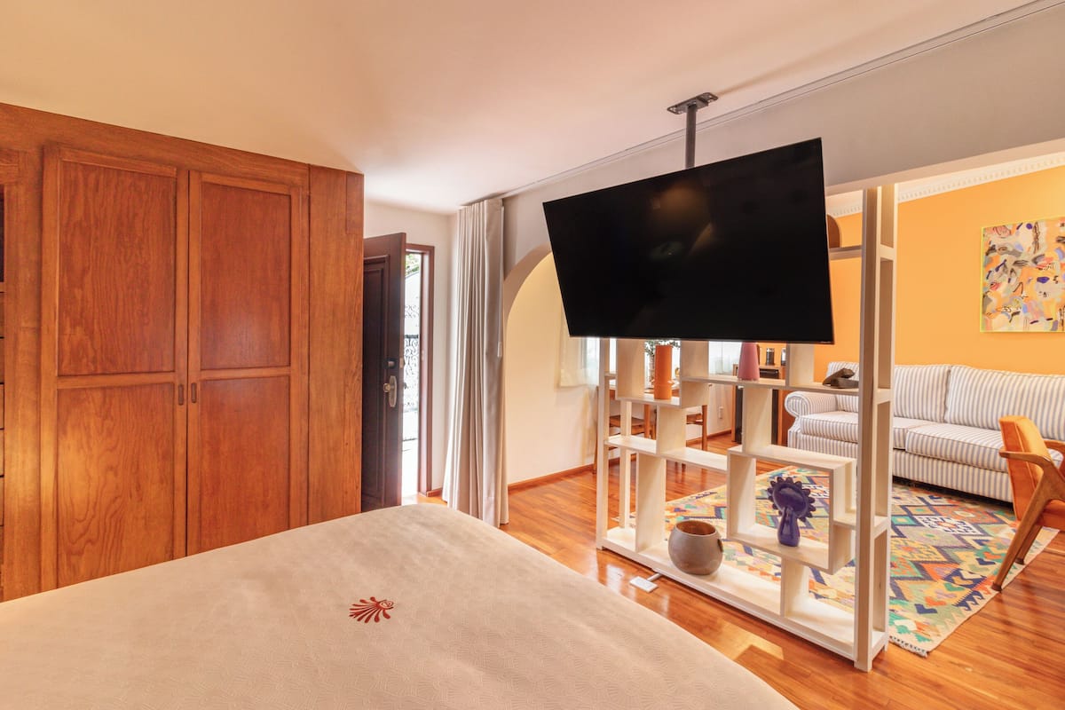 A bedroom with a bed, wooden wardrobe, TV, and a view of a living area with a sofa and a patterned rug.