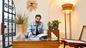 Ein Mann sitzt an einem Holztisch mit Glastischplatte und liest ein Buch in einem hell erleuchteten Raum mit moderner Lampe, Topfpflanzen und Wanddeko.