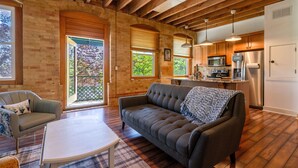 A cozy living room with a sofa, a coffee table, and a fireplace. There is a kitchen area with a refrigerator, microwave, and sink. The room has wooden beams on the ceiling and large windows. - Downtown Grand Haven | Channel Views + Loft (Grand Haven)