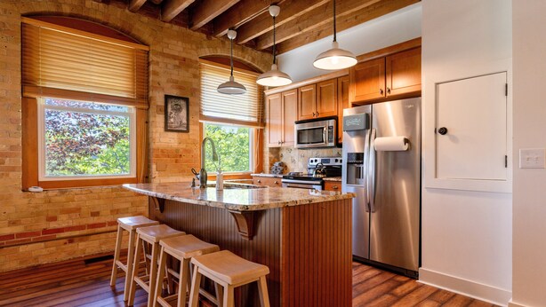 A kitchen with wooden cabinets, a stainless steel refrigerator, a microwave, and a large island with bar stools. - Downtown Grand Haven | Channel Views + Loft (Grand Haven)