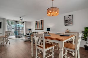Un espace repas avec une table en bois et des chaises blanches, un luminaire suspendu et des œuvres d’art accrochées aux murs.