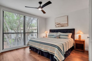 Une chambre à coucher comprenant un lit, deux tables de chevet, un ventilateur de plafond, une fenêtre avec des stores et une œuvre d’art accrochée au mur.