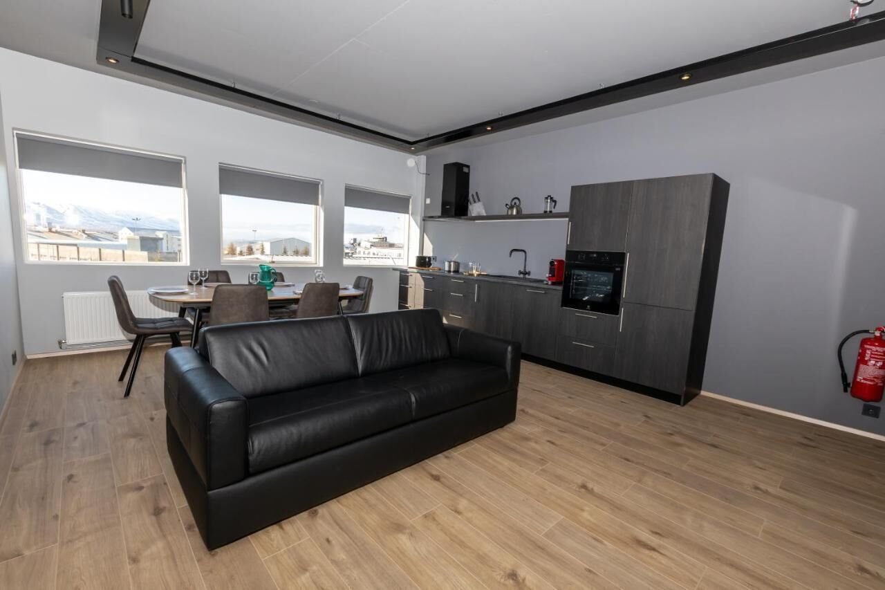 A modern living room with a black leather sofa, a dining area with a table and chairs, and a kitchen with dark cabinetry and a built-in oven.