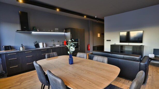 Una cucina moderna con mobili scuri, un tavolo da pranzo in legno e un angolo salotto con televisione a schermo piatto.