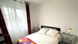 A neatly made bed with white linens and a colorful quilt, two bedside lamps, a window with sheer curtains, and a tiled floor.