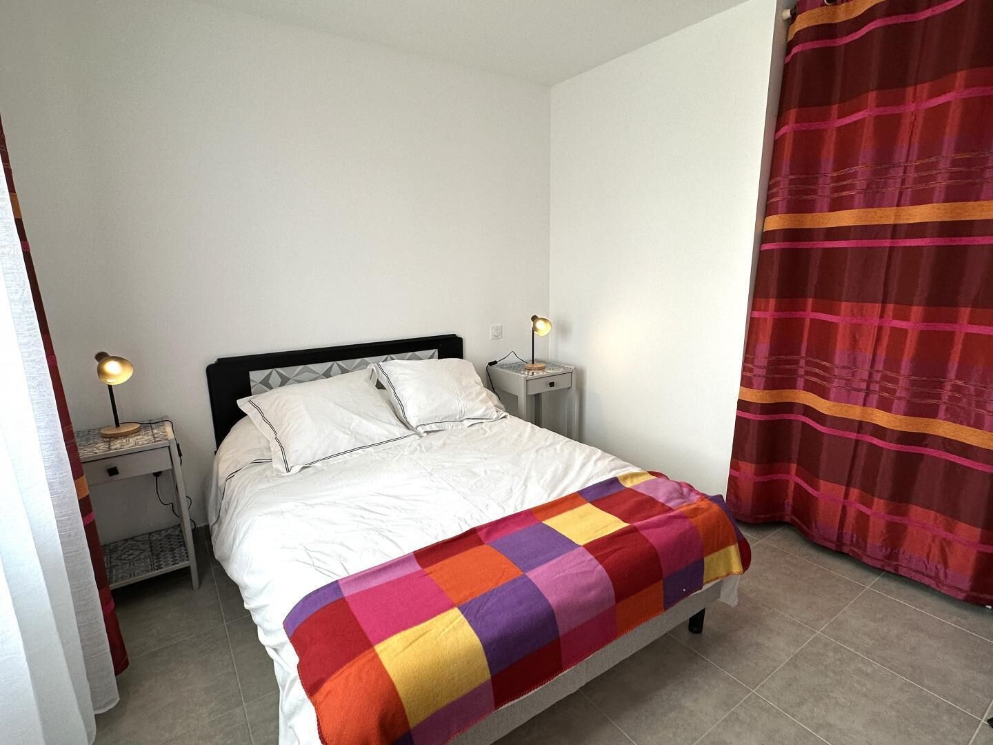 A bed with a white comforter and pillows, a colorful checkered blanket, two bedside lamps, a nightstand, and a red curtain.