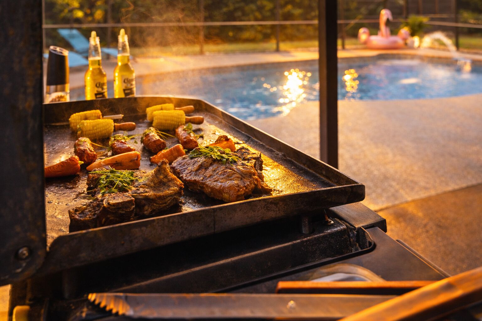 En grill med kött och grönsaker, en pool i bakgrunden och ölflaskor.