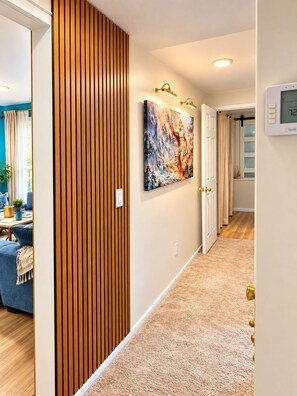 A hallway with a wooden panel wall, a painting, a thermostat, and a glimpse of a living area with a sofa and curtains.