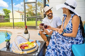 A couple seated by a poolside table with a wine bottle, glasses, and a plate of snacks.