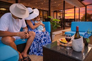 Una pareja sentada en un sofá azul al aire libre, con una mesa delantera que contiene una botella de vino, copas y un plato de comida.
