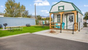 A small, single-story building with a green roof and white walls, featuring a porch with a bench and a table. - Tiny Oasis #1 - Hurricane, UT (Hurricane)