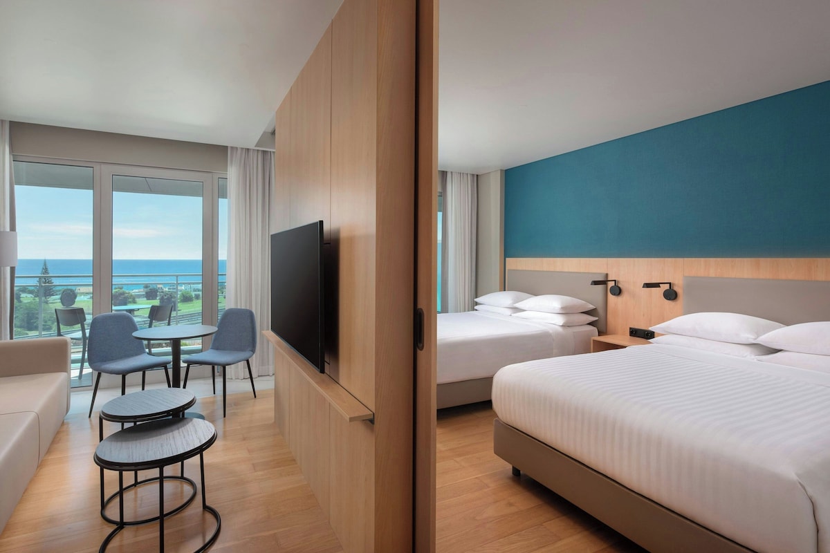 Habitación de hotel con cama, televisor, escritorio y sillas, y vista al océano.