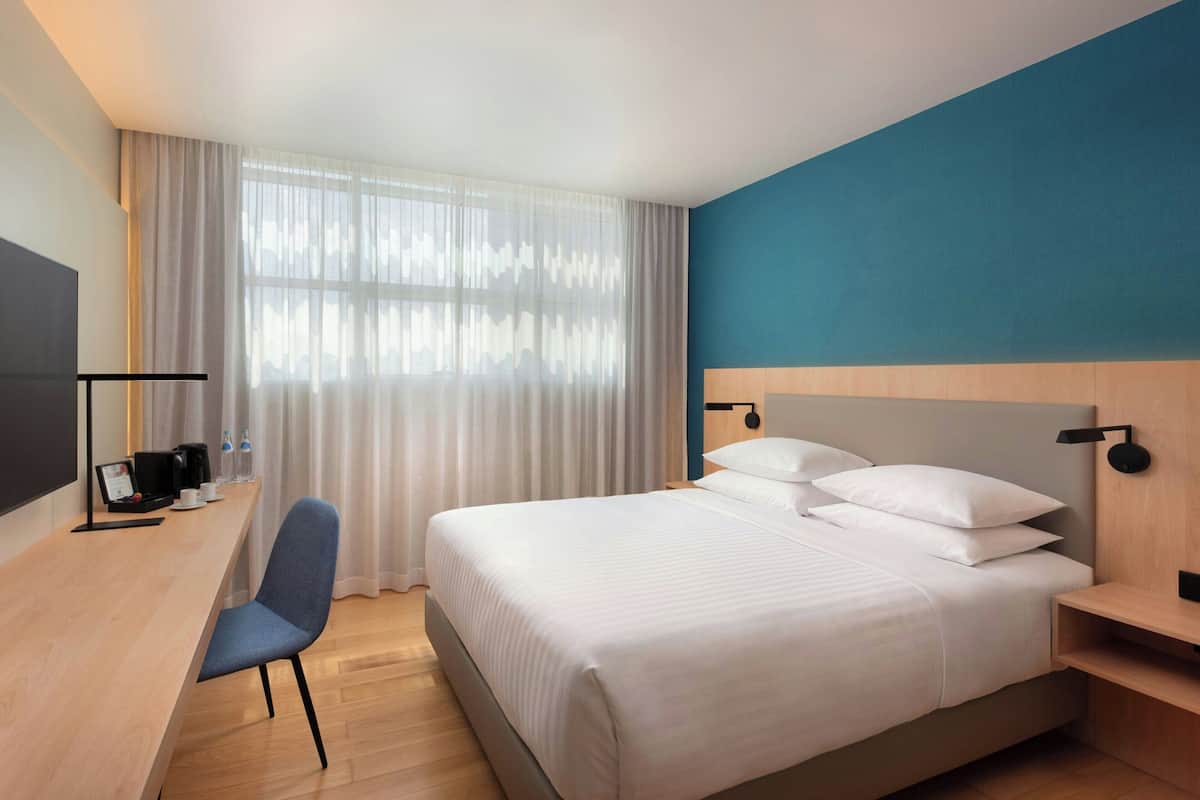 A modern hotel room with a large bed, a desk with a chair, a flat-screen TV, and a window with sheer curtains.