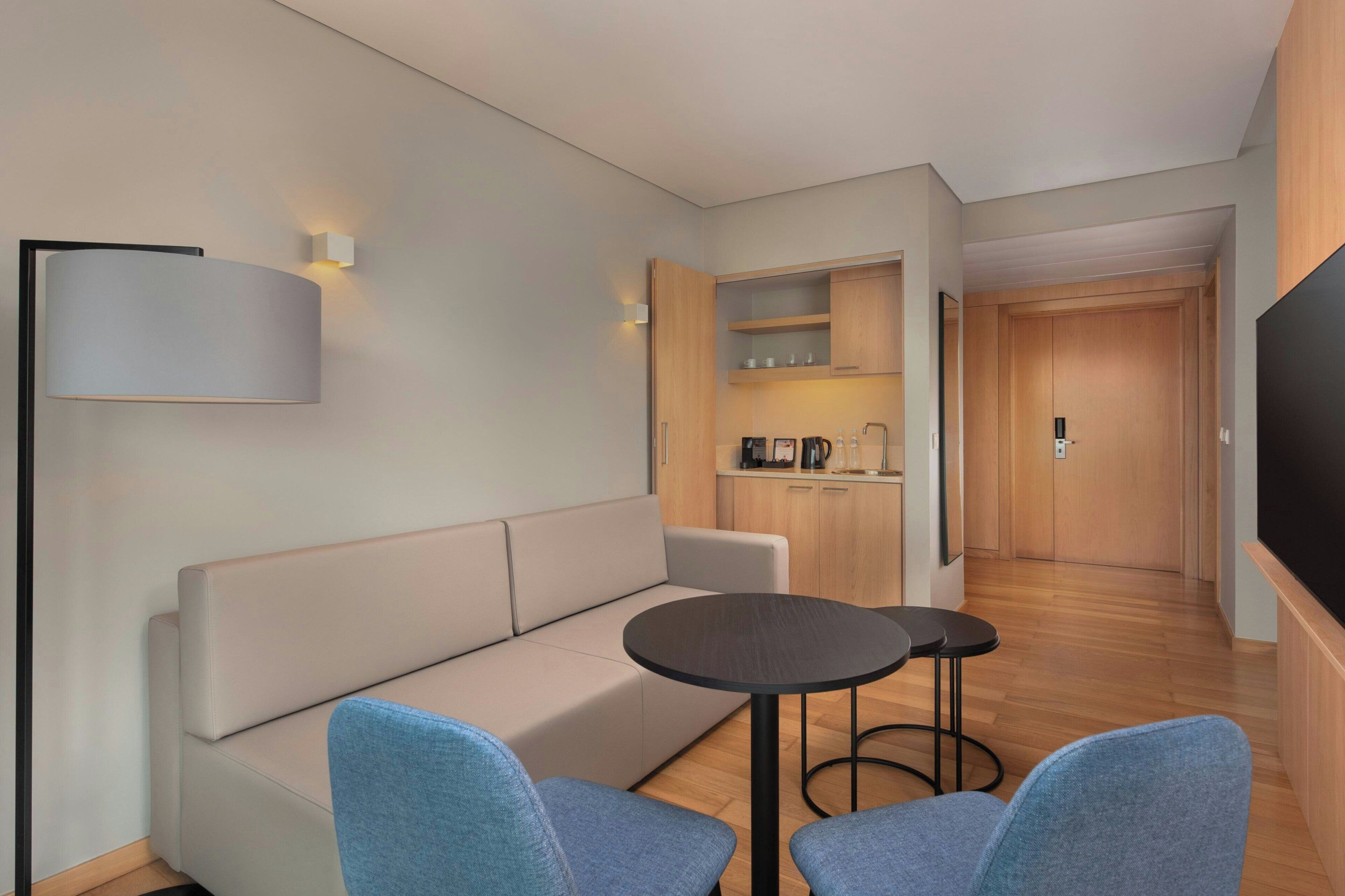A modern hotel room with a sofa, two armchairs, a small round table, a flat-screen TV, a kitchen area with cabinets, and a wooden floor.