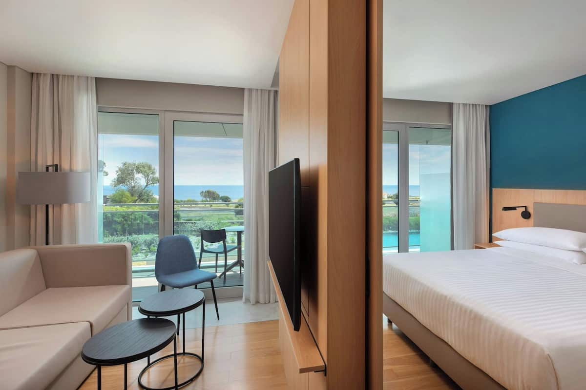 A hotel room with a bed, a sofa, a TV, a small table, and a chair. There is a balcony with a view of the sea.