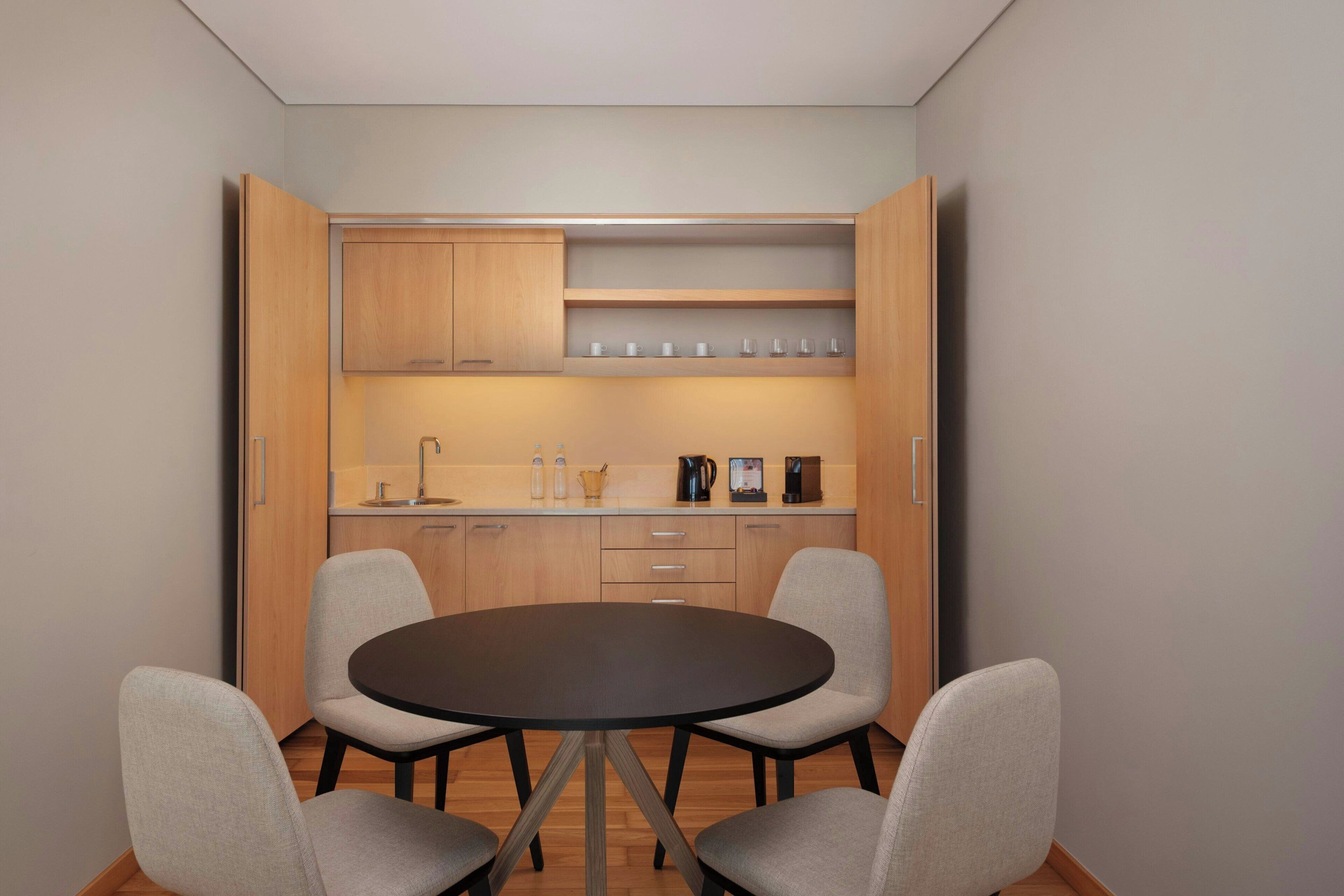 Cozinha compacta com mesa redonda para refeições e quatro cadeiras, armários de madeira e bancada com diversos eletrodomésticos e utensílios.