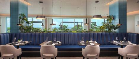 A modern restaurant with a blue booth seating area, multiple tables with chairs, and hanging pendant lights.
