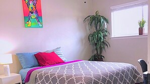 A bedroom with a bed, a ceiling fan, a window with blinds, a potted plant, and a colorful painting on the wall.