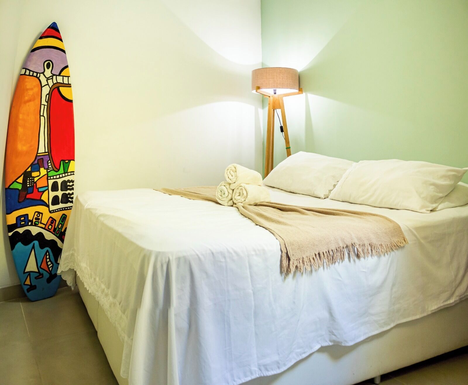 Uma cama com roupa de cama branca e um cobertor bege, uma prancha de surf colorida encostada na parede e um abajur com cúpula bege.