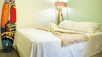 Uma cama com roupa de cama branca e um cobertor bege, uma prancha de surf colorida encostada na parede e um abajur com cúpula bege.