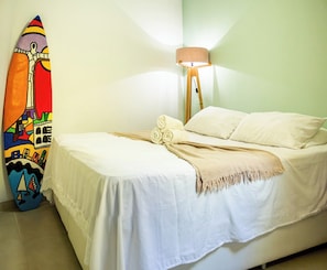 Un lit avec du linge de lit blanc et un plaid beige, une planche de surf colorée appuyée contre le mur et une lampe de sol avec un abat-jour beige.