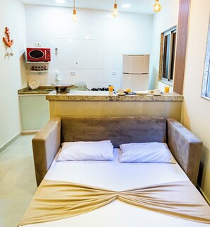 A compact kitchenette with a microwave, stove, and refrigerator. A bed with a beige blanket and pillows is in the foreground.