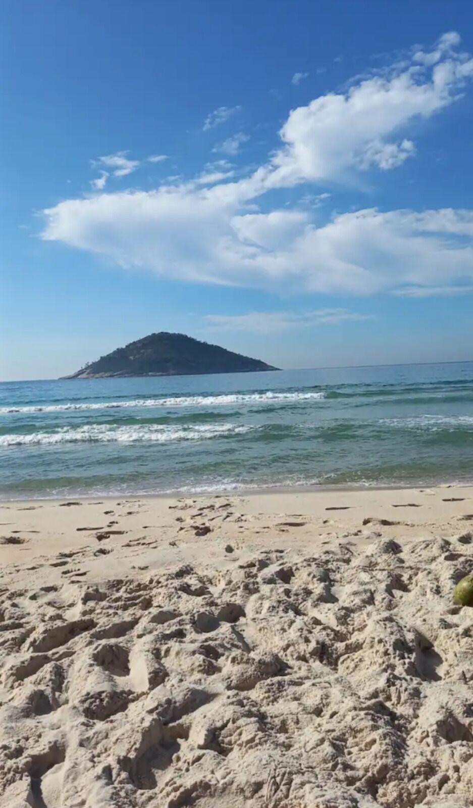 Uma praia com pegadas que levam à água, céu claro e uma ilha ao longe.