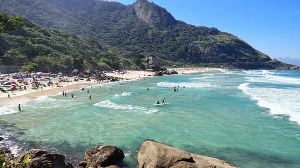 A beach with clear turquoise waters, rocky outcrops, and a mountainous backdrop.