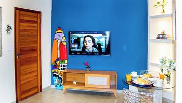 Um quarto com porta de madeira, televisão presa a uma parede azul, um pequeno armário de madeira e uma mesa de vidro com uma cadeira.